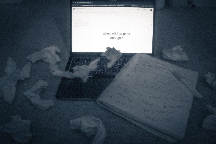 A computer has text that reads "when will i be good enough". There are many crumpled papers surrounding the computer. A notebook lays near the computer open to a page that has negative thoughts and the words "not good enough written on it". The image has been edited to look cold and dark, and the image is blurry and grainy.